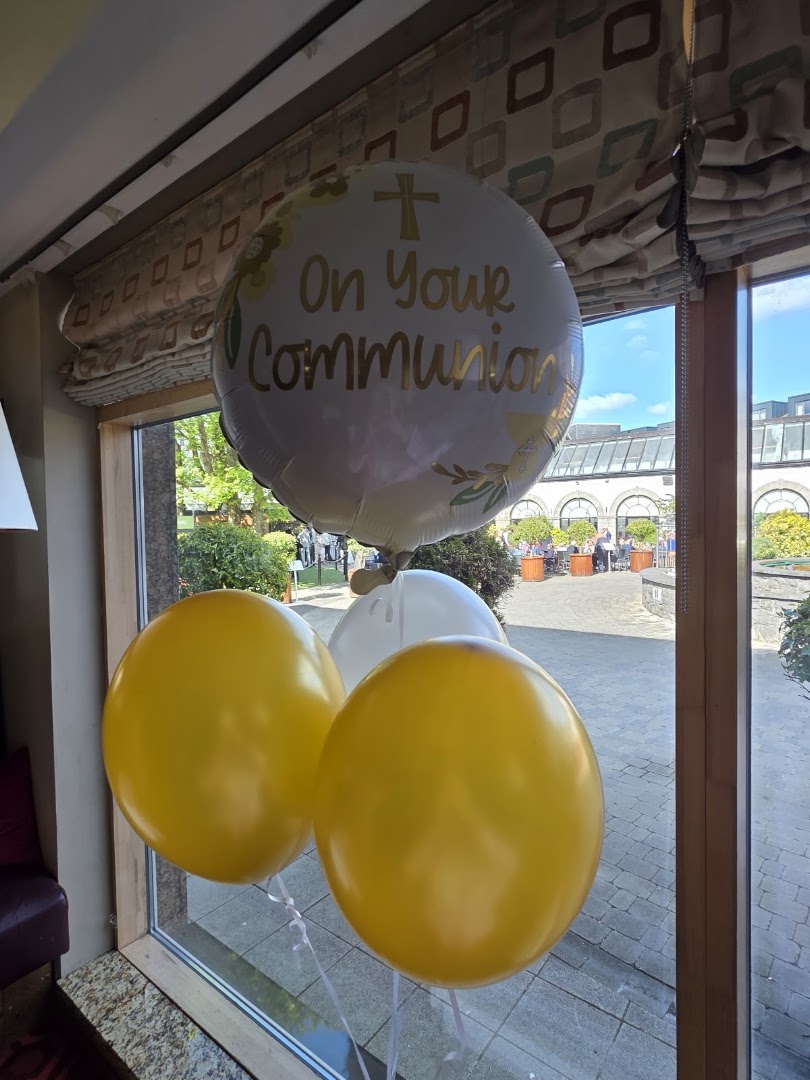 A bunch of balloons including one with 'On Your Communion' text, placed near a window.