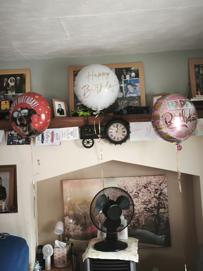 Three birthday balloons floating in a room with a fan and pictures in the background.